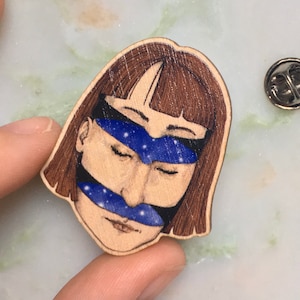 Op de afbeelding: Een houten pin met een handgeschilderde illustratie van een vrouwen gezicht met een blauwe en witte sterrenhemel die haar ogen en mond bedekt.