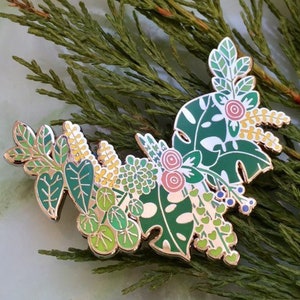 May include: A green, yellow, and pink enamel pin featuring a cluster of stylized leaves and flowers. The pin is designed to look like a bouquet of foliage.