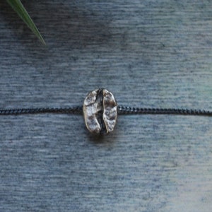 Può includere: Collana con pendente a forma di chicco di caffè argentato. Il pendente presenta linee strutturate, sospeso a una delicata catena scura. Lo sfondo è un blu-grigio tenue con una pianta verde in alto a sinistra.