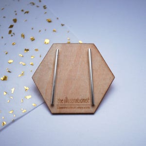May include: A pair of silver bar earrings displayed on a hexagonal wooden stand. The stand has the text "the illustrationist" and "illustrationist.etsy.com" printed on it.