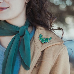 May include: A woman wearing a tan jacket with a green scarf tied around her neck. She is wearing a green floral enamel pin on the lapel of her jacket.