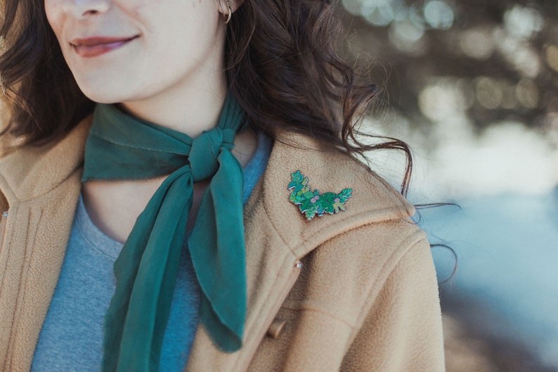 May include: A woman wearing a tan jacket with a green scarf tied around her neck. She is wearing a green floral enamel pin on the lapel of her jacket.