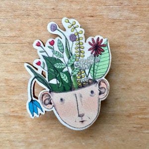 Könnte beinhalten: Eine Holz-Anstecknadel mit einer handgemalten Illustration eines Kopfes, aus dem Blumen wachsen. Der Kopf hat einen hellbraunen Hautton, braune Augen und einen kleinen Mund. Die Blumen sind in verschiedenen Farben, darunter Rot, Rosa, Blau und Gelb.