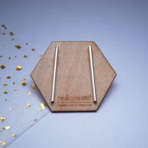 May include: A pair of silver bar earrings displayed on a wooden hexagonal stand. The stand has the text "the illustrationist" and "illustrationist.etsy.com" printed on it.