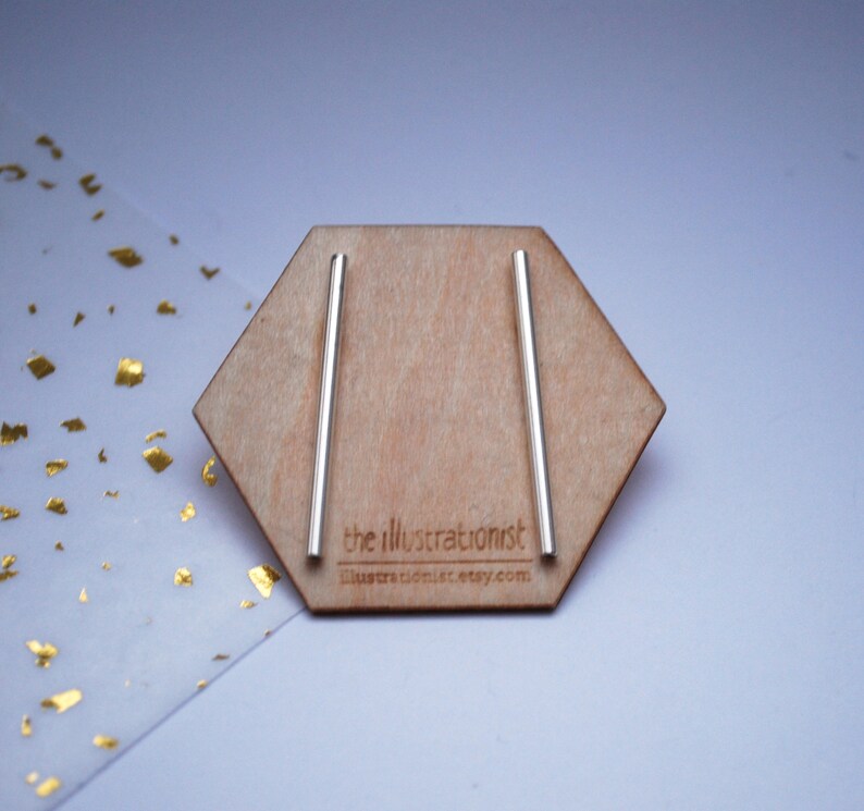 May include: A pair of silver bar earrings displayed on a wooden hexagonal stand. The stand has the text "the illustrationist" and "illustrationist.etsy.com" printed on it.