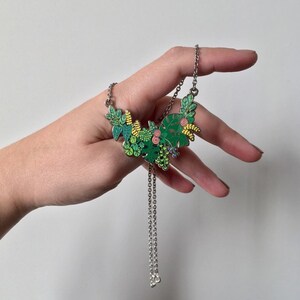 May include: A silver chain necklace with a green, yellow, and pink enamel pendant featuring a floral design. The pendant is shaped like a crescent moon and features various types of leaves and flowers.
