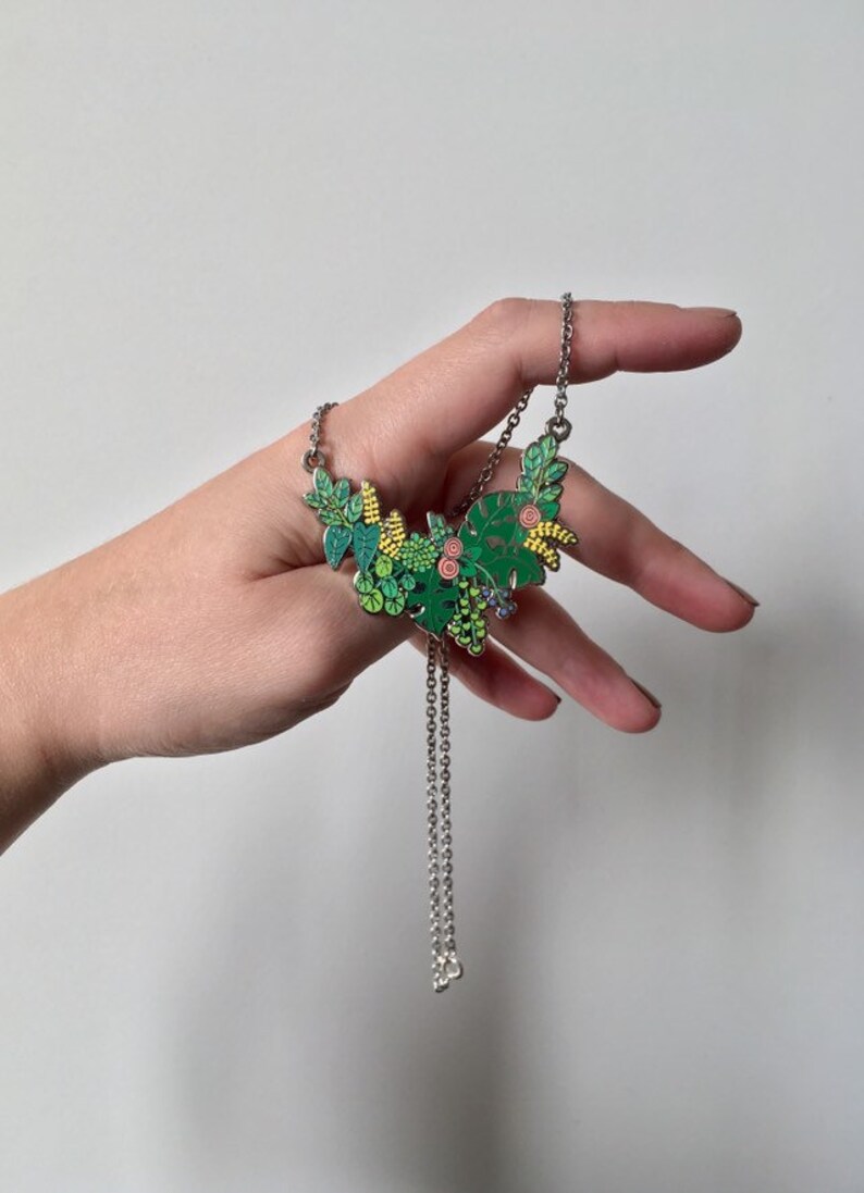 May include: A silver chain necklace with a green, yellow, and pink enamel pendant featuring a floral design. The pendant is shaped like a crescent moon and features various types of leaves and flowers.