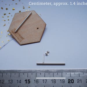 May include: A pair of silver bar earrings, measuring 1.4 inches in length. The earrings are shown on a wooden surface with a ruler for size reference. The ruler shows measurements in centimeters.