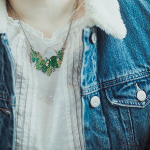 May include: A silver chain necklace with a green and pink enamel pendant shaped like a cluster of leaves. The necklace is worn over a white blouse and a denim jacket.