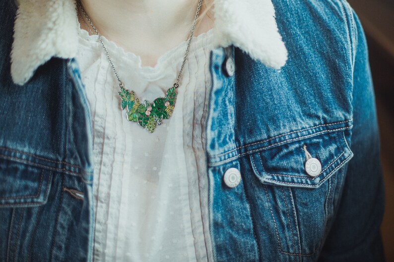 May include: A silver chain necklace with a green and pink enamel pendant shaped like a cluster of leaves. The necklace is worn over a white blouse and a denim jacket.