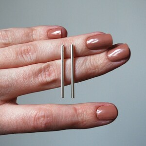 May include: A pair of silver bar earrings, held between two fingers with nude nail polish.
