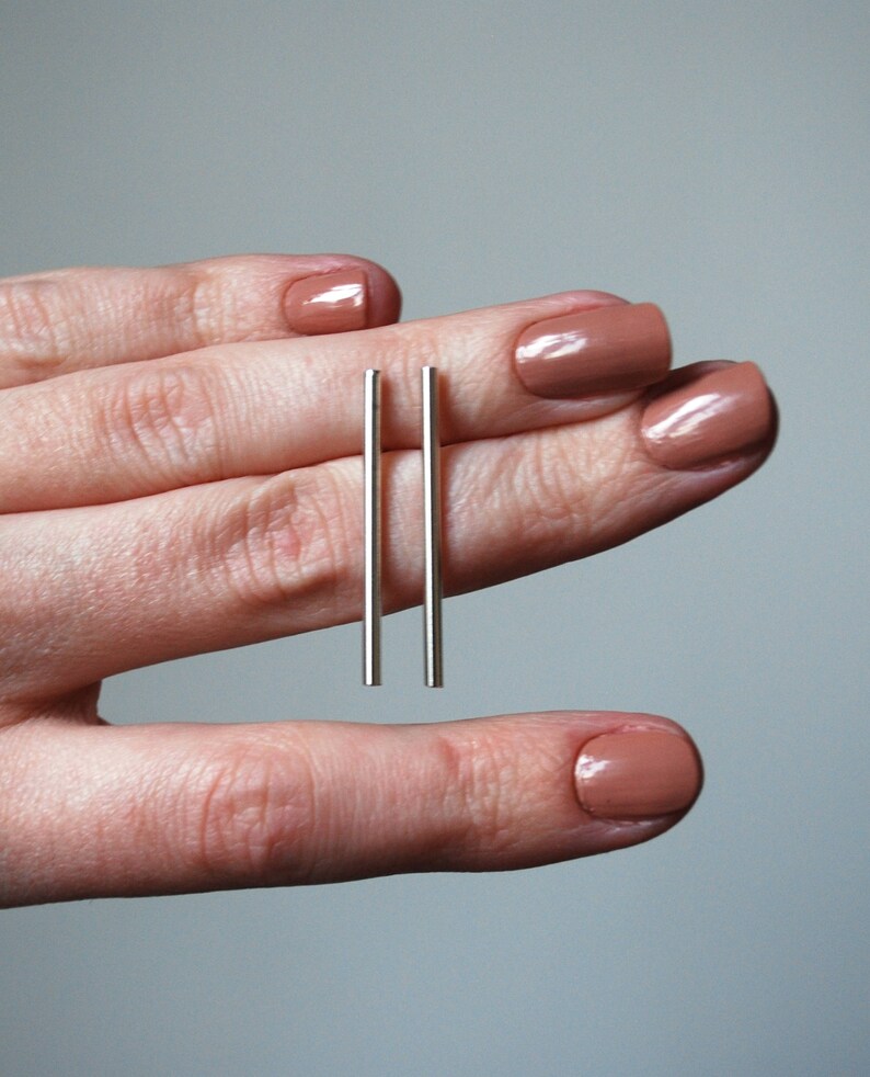 May include: A pair of silver bar earrings, held between two fingers with nude nail polish.