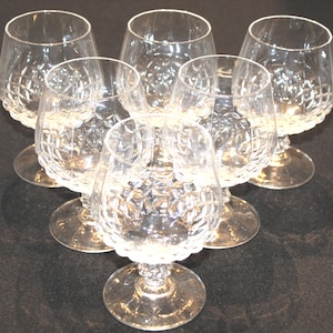 May include: Six clear crystal brandy snifters arranged in a circular pattern. Each glass features a rounded bowl with a diamond-cut pattern and a short stem. The glasses are on a dark surface.