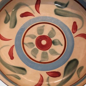 May include: A round ceramic bowl with a cream-colored base featuring a central red circle surrounded by green leaf-like brushstrokes. A wide light blue band encircles the center, accented by thin red rings and scattered red and green painted motifs.