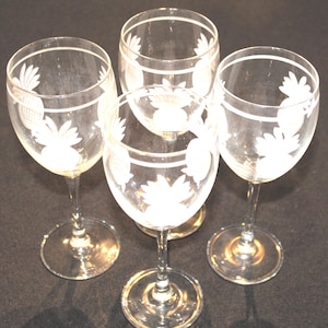 May include: Four clear wine glasses with etched pineapple designs. The glasses have a long stem and a round base. The pineapple design is etched in white on the bowl of the glass. The glasses are arranged on a dark surface.