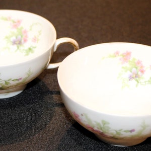 May include: Two white teacups with floral designs. The cups have a delicate floral pattern in shades of pink, purple, and green.