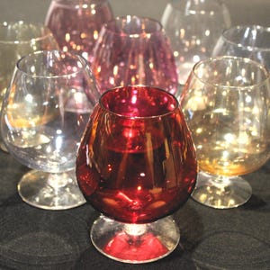 May include: A collection of brandy snifters in various colors, including clear, amber, and ruby red. The glasses have a rounded bowl shape and sit on a short stem with a circular base. The arrangement suggests a display of glassware.