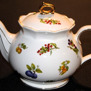 May include: White porcelain teapot with a gold trim and a gold knob on the lid. The teapot is decorated with a floral pattern of red berries, green leaves, and yellow and purple fruit.