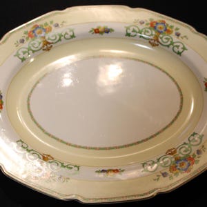 May include: An oval serving platter with a cream-colored background and a decorative border of green, gold, and floral designs. The border features a repeating pattern of stylized flowers and leaves.