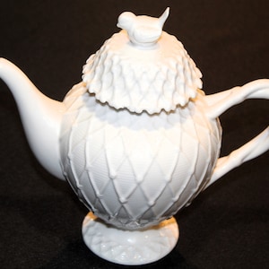 May include: White porcelain teapot with a bird on the lid and a lattice design on the body. The teapot has a round body and a curved handle.