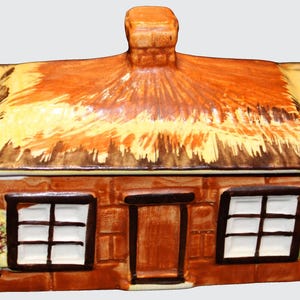 May include: A ceramic cottage-shaped box with a removable lid featuring a textured brown and yellow thatched roof and a central chimney. The base depicts a brown brick house with two white-paned windows, a central door, and floral accents.
