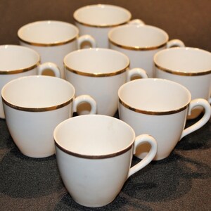 May include: A set of ten white ceramic teacups with gold trim. The cups are arranged in a circle on a dark surface.