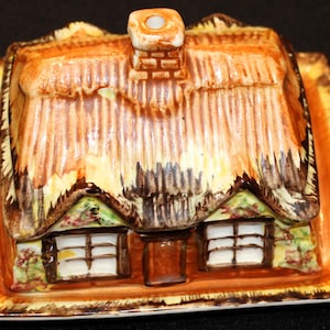 May include: A ceramic cottage-shaped butter dish with a thatched roof and a brown and green painted exterior. The dish is on a matching plate with a gold trim.