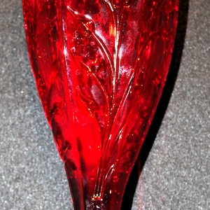 May include: A tall, ruby-red glass vase with a flared base and a slender stem. The vase features an embossed floral design and a textured surface. The vase is approximately 8 inches tall.