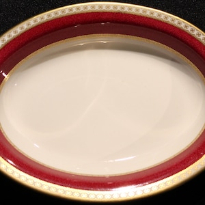 May include: Oval serving dish with a cream-colored interior and a wide, burgundy-colored border. The border is accented with a gold band and a delicate, repeating pattern. The dish is likely made of ceramic or porcelain.