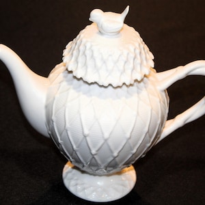 May include: White ceramic teapot with a bird-shaped lid finial. The teapot features a textured, diamond-patterned body and a curved handle. The lid has a ruffled edge. The spout is curved and elegant.