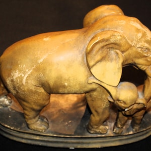 May include: A vintage, tan-colored elephant figurine with a mother elephant and two calves. The sculpture has a weathered appearance, with visible wear and tear. The base is a dark, oval shape. The elephant's trunk is raised, and the calves are nestled close.