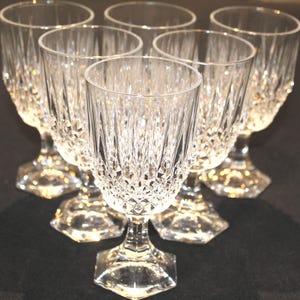 May include: A set of six clear crystal wine glasses. Each glass features a diamond-cut pattern on the bowl and a faceted stem. The glasses are arranged in a circular pattern, showcasing their elegant design.
