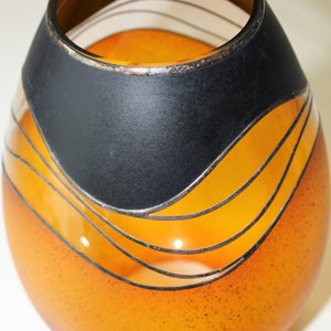 May include: An orange glass votive candle holder with a black band and silver accents. The candle holder has a rounded shape and features horizontal wavy lines. The top rim is dark brown.