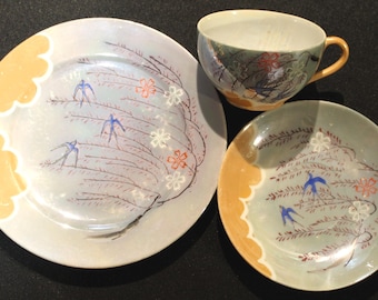 May include: A three-piece ceramic tea set featuring a plate, saucer, and teacup. The set has a light blue background with a design of birds, flowers, and branches. The rims are trimmed with a yellow scalloped edge.