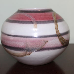 May include: A round ceramic vase with horizontal stripes in shades of white, pink, and dark gray. The vase has a smooth, glossy finish and a small opening at the top. A brown, abstract design is painted on the side.