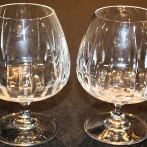 May include: Two clear, stemmed brandy snifters with a rounded bowl and a wide base. The glass has a vertical, ribbed pattern. The glasses are empty and sit on a dark surface.
