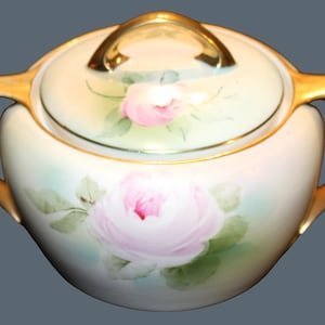 May include: A porcelain sugar bowl with a lid, featuring hand-painted pink roses and green leaves on a cream-colored background. The bowl has two gold-toned handles and a gold-toned lid handle, with gold trim along the edges.