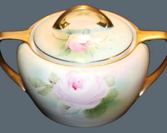 May include: A porcelain sugar bowl with a lid, featuring hand-painted pink roses and green leaves on a cream-colored background. The bowl has two gold-toned handles and a gold-toned lid handle, with gold trim along the edges.