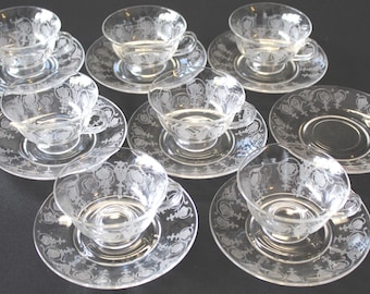 May include: A set of clear glass teacups and saucers. Each cup and saucer features an etched floral design. The cups have handles and sit on matching saucers. The set includes eight cups and saucers, perfect for serving tea.