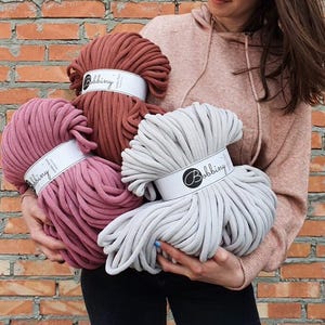 Puede incluir: Tres ovillos de hilo de cuerda de algodón grueso en varios colores: terracota, rosa polvoriento y gris claro. Cada ovillo tiene una etiqueta blanca con el texto "Bobbiny" impreso en ella.