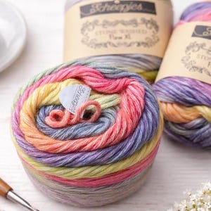 Op de afbeelding: Meerkleurige Scheepjes-garenballen in roze, geel en paars. Een haaknaald en een kopje koffie zijn ook zichtbaar, wat wijst op een knutselthema. Het garen is gemarkeerd met "Stone Washed Flow XL".