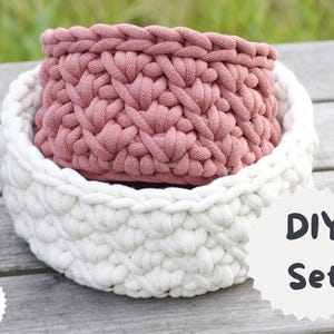 Puede incluir: Dos cestas de ganchillo hechas a mano. La cesta más grande es blanca y la más pequeña del interior es rosa. Las cestas están hechas de hilo grueso y tienen un aspecto texturizado. La imagen también incluye el texto "DIY Set".