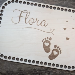 May include: A light-colored wooden plaque with the name "Flora" in cursive script. The plaque features engraved baby footprints with heart cutouts, small heart shapes, and a decorative border of evenly spaced holes.