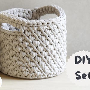 Puede incluir: Una cesta de ganchillo hecha a mano, de color gris claro, con dos asas. La cesta tiene un diseño tejido texturizado y es de forma cilíndrica. Las palabras "DIY Set" están en una pegatina blanca junto a la cesta.
