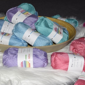 Könnte beinhalten: Eine Schüssel gefüllt mit verschiedenen Farben von Garn, darunter hellblau, lila und rosa. Das Garn ist mit "Carina" gekennzeichnet und hat 50 Gramm pro Knäuel.
