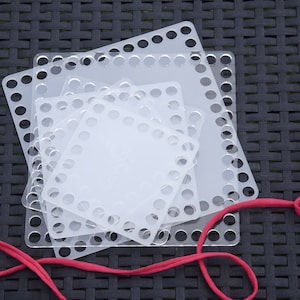 May include: Four square plastic loom boards with holes for weaving yarn. The boards are stacked on top of each other and a red cord is wrapped around them.