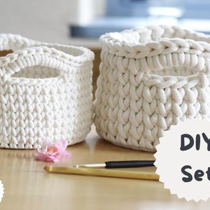 Puede incluir: Dos cestas de ganchillo hechas a mano, de color blanco roto, con aberturas para las asas. Las cestas están hechas de hilo grueso y texturizado. La imagen también incluye ganchos de ganchillo y el texto "DIY Set". Un proyecto de manualidades.
