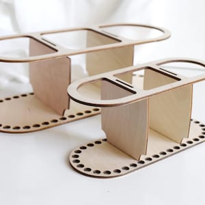 Könnte beinhalten: Zwei Holz-Organizer mit natürlichem Finish. Jeder hat eine ovale Basis mit gleichmäßig verteilten Löchern, vertikalen Stützen und einem oberen Abschnitt mit drei Öffnungen. Das Design ist schlicht und modern.
