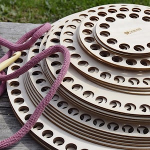 Può includere: Un set di telai circolari per la maglia in legno di diverse dimensioni, impilati uno sopra l'altro. I telai hanno dei fori attorno ai bordi e sono fatti di legno chiaro. Un cordoncino da uncinetto rosa e un uncinetto di legno sono accanto ai telai.