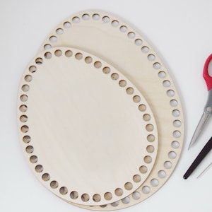 Op de afbeelding: Twee ovale houten weefringen, een kleiner dan de andere, met gaten rond de randen. Ook inbegrepen zijn een rode schaar, een haaknaald en een tapijtnaald.